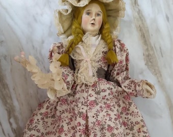 May include: A vintage porcelain doll with blonde hair in braids, wearing a pink and white floral dress, a white lace bonnet with a pink bow, and black shoes with white stripes.