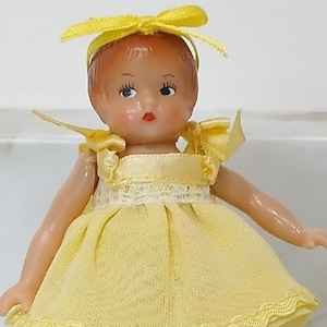 May include: A small doll with reddish-brown hair styled with a yellow ribbon, wearing a sleeveless yellow dress with a white lace trim at the waist and a scalloped hem. The doll has white socks and black shoes, and is supported by a white stand.