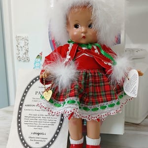 May include: A vintage Patsy doll wearing a red and green plaid dress with white lace trim, a matching jacket, and a white feathered hat. The doll has red shoes and a gold heart-shaped charm. A certificate of authenticity is included.