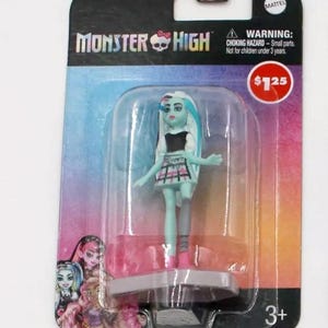 May include: A Monster High doll in a clear plastic package. The doll has blue skin, white and blue hair, and is wearing a black top and plaid skirt. The packaging includes the Monster High logo and a warning about small parts.