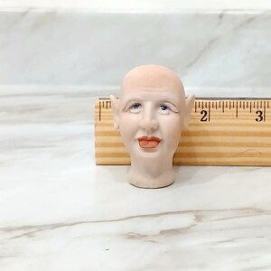 May include: A pale, bald doll head with pointed ears, blue eyes, and orange-toned lips, positioned in front of a wooden ruler showing a width of approximately 1.5 inches for the head, with the ruler markings clearly visible behind the object.