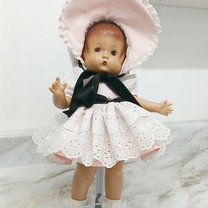 May include: A doll with brown hair wearing a light pink bonnet with white trim, a white short-sleeved dress with a layered eyelet lace skirt, a black ribbon tie at the neck, white ruffled socks, and black shoes, supported by a white metal stand.
