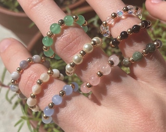 Peut inclure: Un ensemble de six bagues en perles, chacune avec une couleur et un type de perle différents. Les bagues sont empilées sur un doigt, la bague verte en haut et la bague bleue en bas.