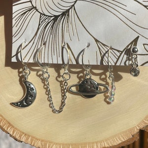 May include: A set of five silver hoop earrings with celestial charms. The charms include a crescent moon, a chain, a planet with rings, and a strand of iridescent beads.
