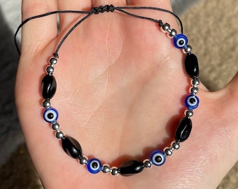 May include: A black string bracelet with silver and blue beads. The beads are in the shape of evil eyes.
