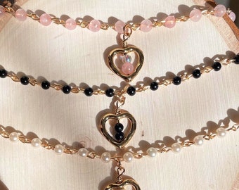 Peut inclure: Trois pendentifs en forme de cœur dorés avec des perles de différentes couleurs à l'intérieur. Le pendentif du haut a des perles roses, le pendentif du milieu a des perles noires et le pendentif du bas a des perles blanches. Les pendentifs sont suspendus à des chaînes avec des perles roses, noires et blanches.