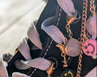 Könnte beinhalten: Nahaufnahme eines schwarzen Canvas-Schuhs mit weißen Schnürsenkeln, verziert mit verschiedenen goldfarbenen Anhängern. Zu den Anhängern gehören ein Herz, ein Smiley, ein rosa Getränkedosen-Tab und ein Anhänger mit der Aufschrift "Happy".
