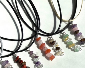 Puede incluir: Cinco collares con colgantes de piedras preciosas de diferentes colores. Cada colgante está hecho de pequeñas piedras de forma irregular. Los collares están sobre una superficie blanca.