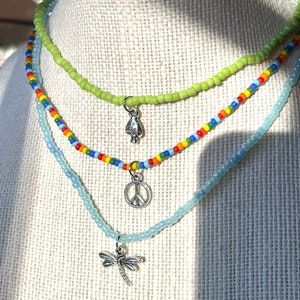 Puede incluir: Tres collares de cuentas superpuestos con dijes de plata. El collar superior es verde claro, el collar del medio es de colores arco&iacute;ris y el collar inferior es azul claro. Los dijes son un hongo, un signo de paz y una lib&eacute;lula.