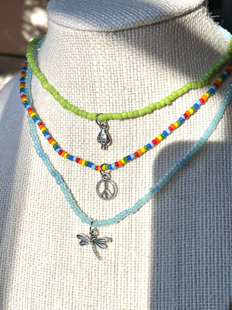 Puede incluir: Tres collares de cuentas superpuestos con dijes de plata. El collar superior es verde claro, el collar del medio es de colores arco&iacute;ris y el collar inferior es azul claro. Los dijes son un hongo, un signo de paz y una lib&eacute;lula.