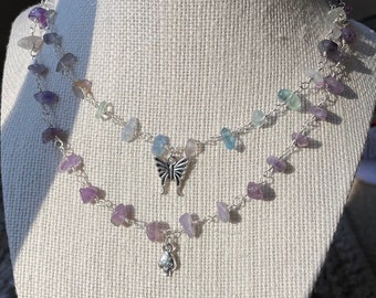 Puede incluir: Un collar de dos capas con una cadena plateada y pequeñas piedras de colores. El collar tiene dos colgantes: una mariposa y un hongo.