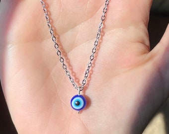 Puede incluir: Un collar de cadena plateada con un dije de ojo turco azul. El dije tiene un centro blanco y una pupila negra.