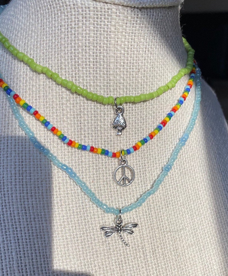 Puede incluir: Tres collares de cuentas superpuestos con dijes. La capa superior es verde claro, la capa intermedia es de colores arco&iacute;ris y la capa inferior es azul claro. Los dijes son un hongo, un signo de paz y una lib&eacute;lula.
