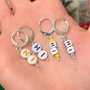 Puede incluir: Tres pares de pendientes de aro de plata con cuentas de letras que deletrean "HI". Las cuentas son blancas con acentos azules y amarillos.