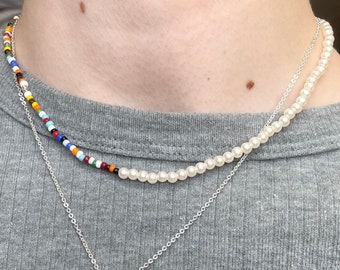 May include: Two necklaces layered on a person's neck. The top necklace is made of small, colourful beads. The bottom necklace is made of white pearls. The bottom necklace has a silver pendant shaped like a sun with rays.