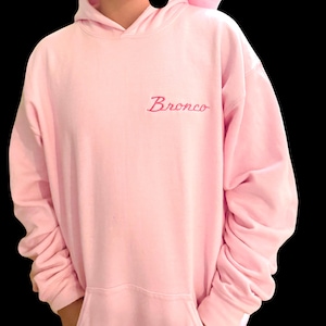 May include: A light pink hoodie with a front pocket and a hood. The word "Bronco" is embroidered in a cursive font on the chest. The sleeves are long and the material appears to be soft.