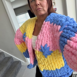 May include: A colourful, chunky knit cardigan with a patchwork design. The sweater features large blocks of pink, yellow, and blue yarn. The sleeves are long and the style is oversized. A cosy and colourful garment.