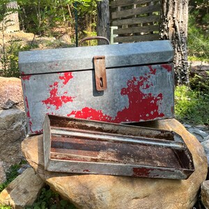 May include: A weathered metal toolbox with red paint accents and a matching tray. The toolbox has a handle and a latch. The items are resting on a large rock, with greenery in the foreground and trees in the background.