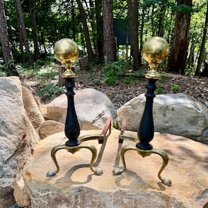 May include: A pair of antique brass and black fireplace andirons. Each andiron features a gold-colored sphere atop a black column, with curved legs. The andirons are displayed on a large rock outdoors.