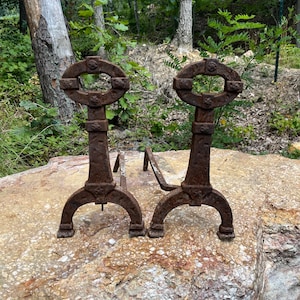 May include: Two antique, rusty metal fireplace andirons. Each andiron has a decorative oval top, a vertical support, and curved legs. The andirons are displayed on a large, textured rock with a natural, outdoor background.