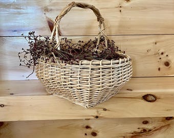 May include: A woven wicker basket with a heart-shaped base and a curved handle. The basket is empty and sitting on a wooden shelf.