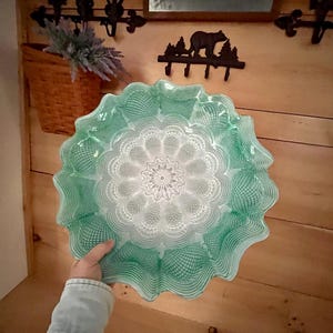 May include: A large, decorative glass plate with a scalloped edge. The plate features a detailed, intricate pattern in shades of green and white, resembling a floral design. The plate is held against a wooden background.