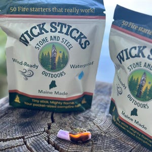 May include: Two packages of Wick Sticks fire starters with the text "50 Fire starters that really work!". The packages are white with a logo of a knife and trees. The text "Wind-Ready" and "Waterproof" are also visible. A lit fire starter is in the foreground.
