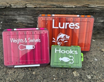 May include: Three clear plastic fishing tackle boxes. The boxes are labeled "Lures", "Weights & Swivels", and "Hooks". The "Lures" box is orange, the "Weights & Swivels" box is pink, and the "Hooks" box is green. Each box has a white label.