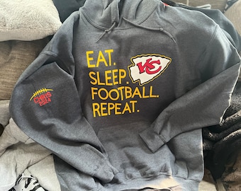 以下が含まれることがあります： グレーのフード付きスウェットシャツに、黄色で「Eat. Sleep. Football. Repeat.」と、赤と白のカンザスシティチーフスのロゴがプリントされています。左袖には、黄色でフットボールと「Chris 2024.」の文字がプリントされています。