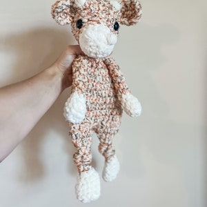 May include: A crocheted brown and white cow plush toy with black eyes. The toy is being held by a hand.