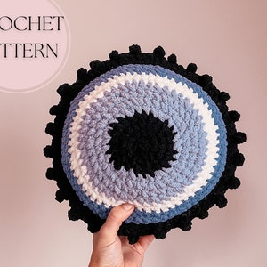 Puede incluir: Patrón de crochet para una almohada en forma de ojo azul, blanco y negro con pompones negros alrededor del borde.