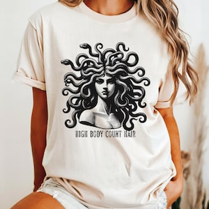 Op de afbeelding: Een crèmekleurig T-shirt met een zwart-witte illustratie van Medusa met de tekst "High Body Count Hair".