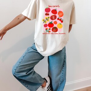 Puede incluir: Una camiseta de color crema con un gráfico de varios tomates rojos, naranjas y amarillos sobre un fondo rectangular rosa. El texto "be cool" está impreso en rojo sobre el gráfico, y "don't be all, like, uncool" está impreso en rojo debajo.