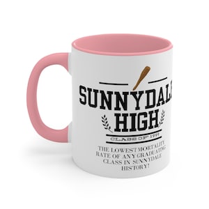 Op de afbeelding: Een witte keramische mok met een roze rand. De mok heeft een zwart-witte afbeelding met de woorden "Sunnydale High" met een honkbalknuppel en de tekst "Class of 1999". De tekst eronder luidt "The lowest mortality rate of any graduating class in Sunnydale history!"
