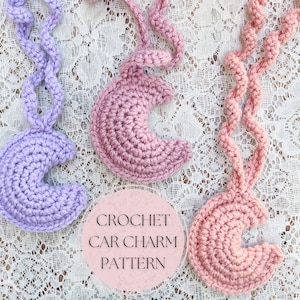May include: Two crocheted car charms in the shape of a crescent moon. One charm is lavender and the other is pink. The charms are hanging from a crocheted chain. The text "CROCHET CAR CHARM PATTERN" is visible in the image.