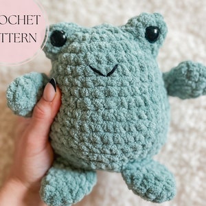 May include: A plush, light teal crochet frog with black button eyes and a stitched smile. The frog has small arms and legs. The text "CROCHET PATTERN" is in a pink circle in the upper left corner.