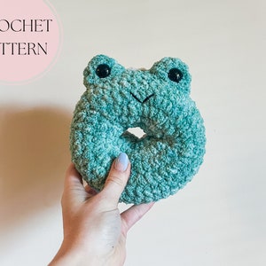 May include: A crocheted frog-shaped donut in a teal green colour. The frog has black eyes and a small, crocheted smile.