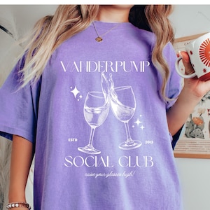 Op de afbeelding: Paars T-shirt met de tekst "Vanderpump Social Club" en een afbeelding van twee wijnglazen die klinken. De tekst "Est. 2013" staat onder de glazen en de tekst "Raise your glasses high!" staat onder de clubnaam.