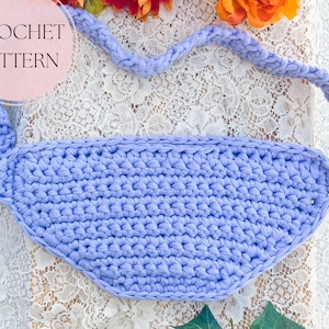 May include: A light purple crocheted bag with a long strap. The bag is lying on a white lace fabric.