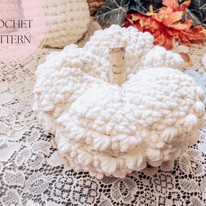 Peut inclure: Citrouille blanche en crochet avec une tige en bois. La citrouille est posée sur une nappe en dentelle blanche. Le texte "CROCHET PATTERN" est visible dans le coin supérieur gauche de l'image.