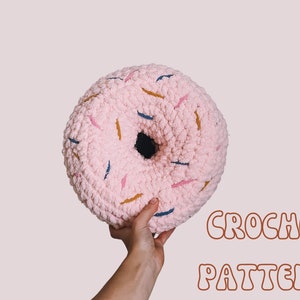 May include: A pink crocheted donut pillow with colorful sprinkles. The pillow is being held by a hand against a light pink background. The text "CROCHET PATTERN" is written in brown letters below the pillow.