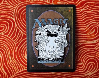 May include: White enamel pin featuring a stylized illustration of a woman's face surrounded by foliage. The pin is set against a Magic: The Gathering card background.