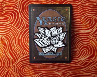 May include: A white enamel pin featuring a stylized black and white illustration of a lotus flower. The pin is on a Magic: The Gathering card with the text "Deckmaster" at the bottom.