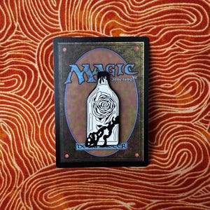 May include: A black and white enamel pin featuring a glass bottle with a rose inside. The bottle is on a black background with the text "Magic Gathering" and "Deck Master" in white.
