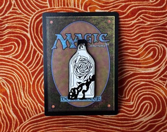 May include: A black and white enamel pin featuring a glass bottle with a rose inside. The bottle is on a black background with the text "Magic Gathering" and "Deck Master" in white.