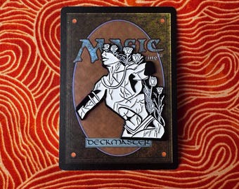 May include: A black and white enamel pin featuring a stylized illustration of a figure with flowers growing from their head. The pin is set against a gold background with the text "Magic" and "Deckmaster" in black.