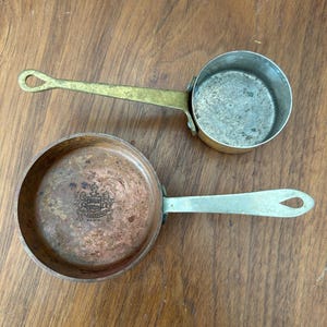 May include: Two small vintage copper and brass saucepans. One saucepan is copper with a brass handle and a decorative emblem. The other is a silver-colored metal with a brass handle. Both have a loop at the end of the handle.