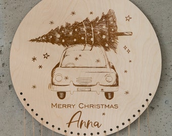 Op de afbeelding: Een cirkelvormige plaat van lichtgekleurd hout met een lasergegraveerde illustratie van een auto met een kerstboom op het dak. De tekst MERRY CHRISTMAS en de naam Anna zijn onder de auto gegraveerd, met gaatjes langs de onderste rand.
