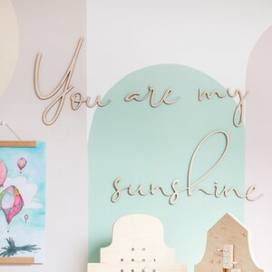 May include: A wooden pegboard activity set with two hinged panels, featuring various small wooden shapes attached to the board. Beside it sits a pastel-coloured stacking ring toy, a watercolour print of hot air balloons, and the wooden text You are my sunshine.