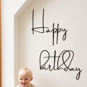 Puede incluir: Un bebé sonriente con un conjunto beige está sentado cerca de una ventana. Encima, letras cursivas negras deletrean "Happy Birthday" en una pared blanca. La escena es brillante y minimalista, perfecta para una celebración de cumpleaños.